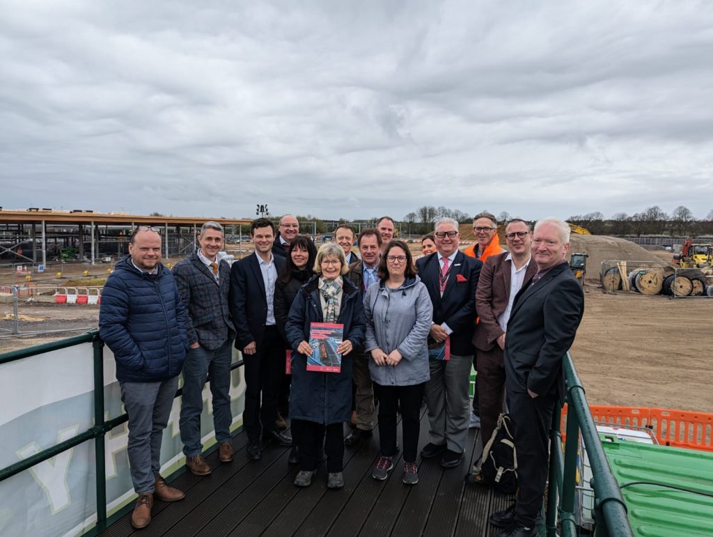 Members of the Transport East Forum and officers at the construction site for the new Beaulieu Park rail station. A group of people standing on a platform at a construction site. The background shows an expansive construction area with various equipment, materials, and partially built structures.