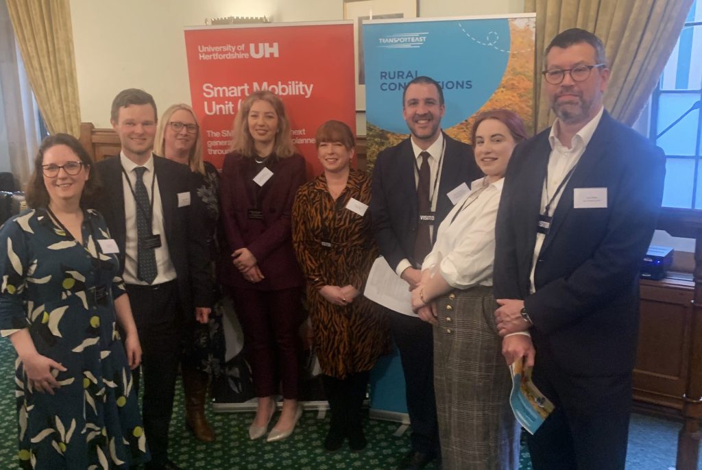 A group of people standing in front of two banners. The left banner is red and reads "University of Hertfordshire UH Smart Mobility Unit." The right banner is blue and reads "Transport East Rural Connections." The individuals are dressed in business attire, with some holding papers or booklets.
