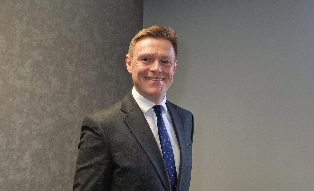 Will Quince, Chair of Transport East. He is wearing a dark grey suit, white shirt, and a navy blue tie with white polka dots and is standing against a grey textured wall.