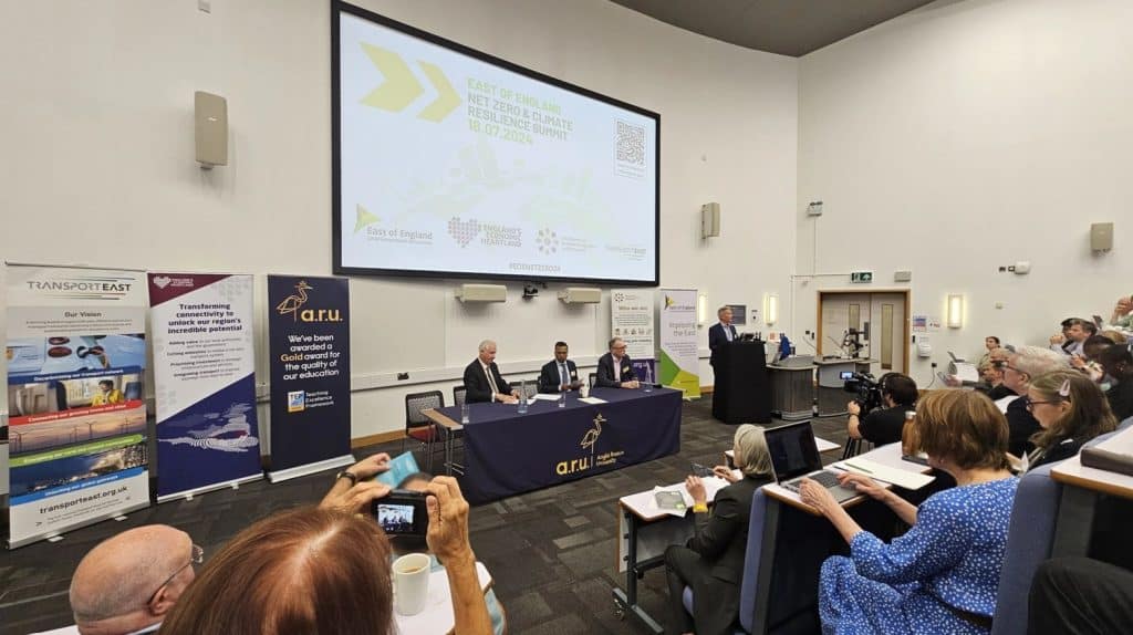 A conference room filled with attendees for the East's Net Zero Event. At the front of the room, four men are seated at a table covered with a blue cloth bearing the logo of Anglia Ruskin University (ARU). Behind them is a large screen displaying information about the "East of England Net Zero & Climate Resilience Summit" scheduled for 18.07.2024. Several banners are positioned to the left and right of the screen, promoting various organizations involved in transportation and education. One man stands at a podium addressing the audience.