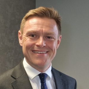 Will Quince, Chair of Transport East. He is wearing a dark grey suit, white shirt, and a navy blue tie with white polka dots and is standing against a grey textured wall.